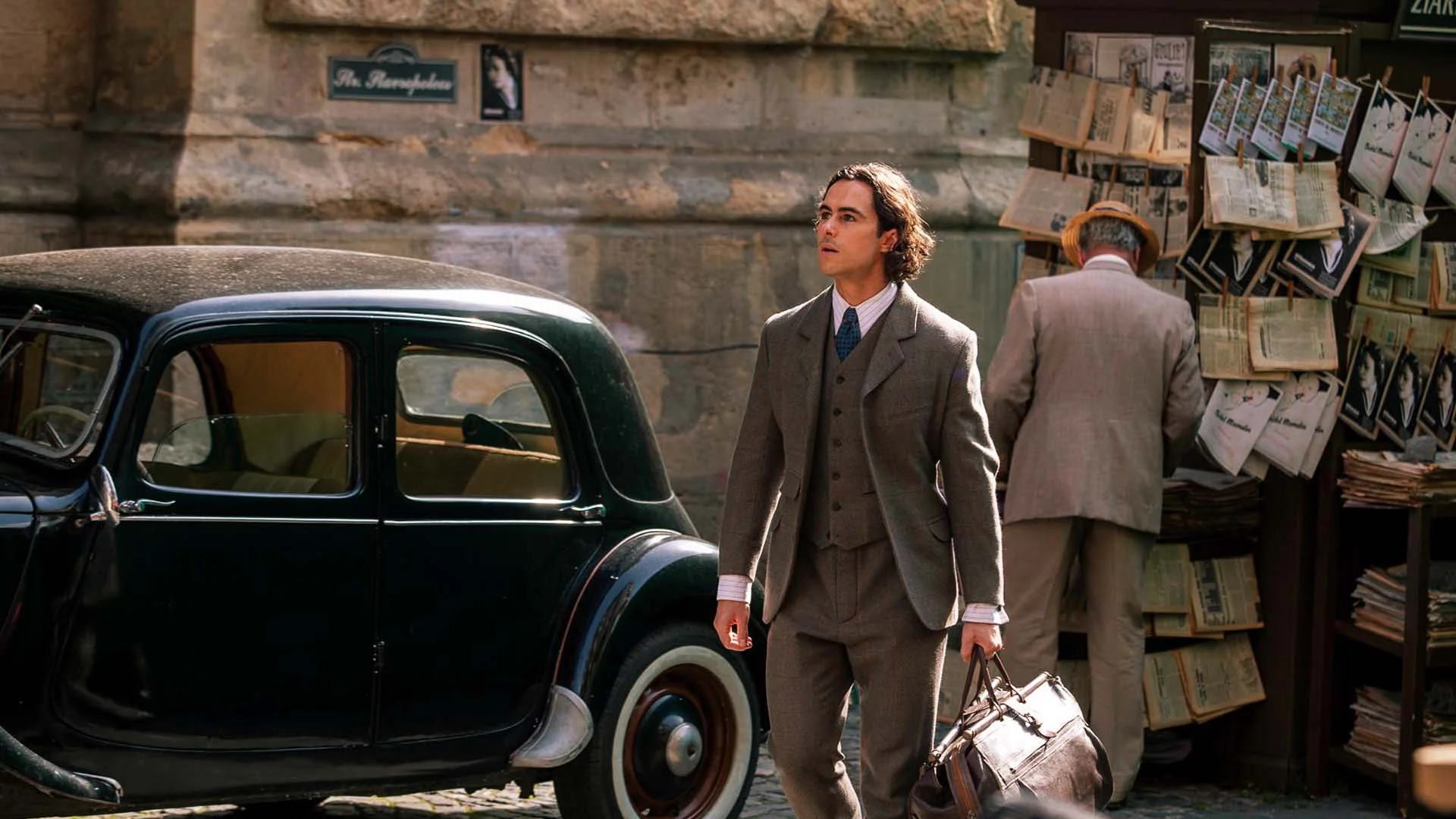 a Man in a 1930s Brown Suit Stands Beside a Dark Vintage Car on a Cobblestone Street Carrying a Leather Bag While Another Man Browses a Newspaper Stand in the Background