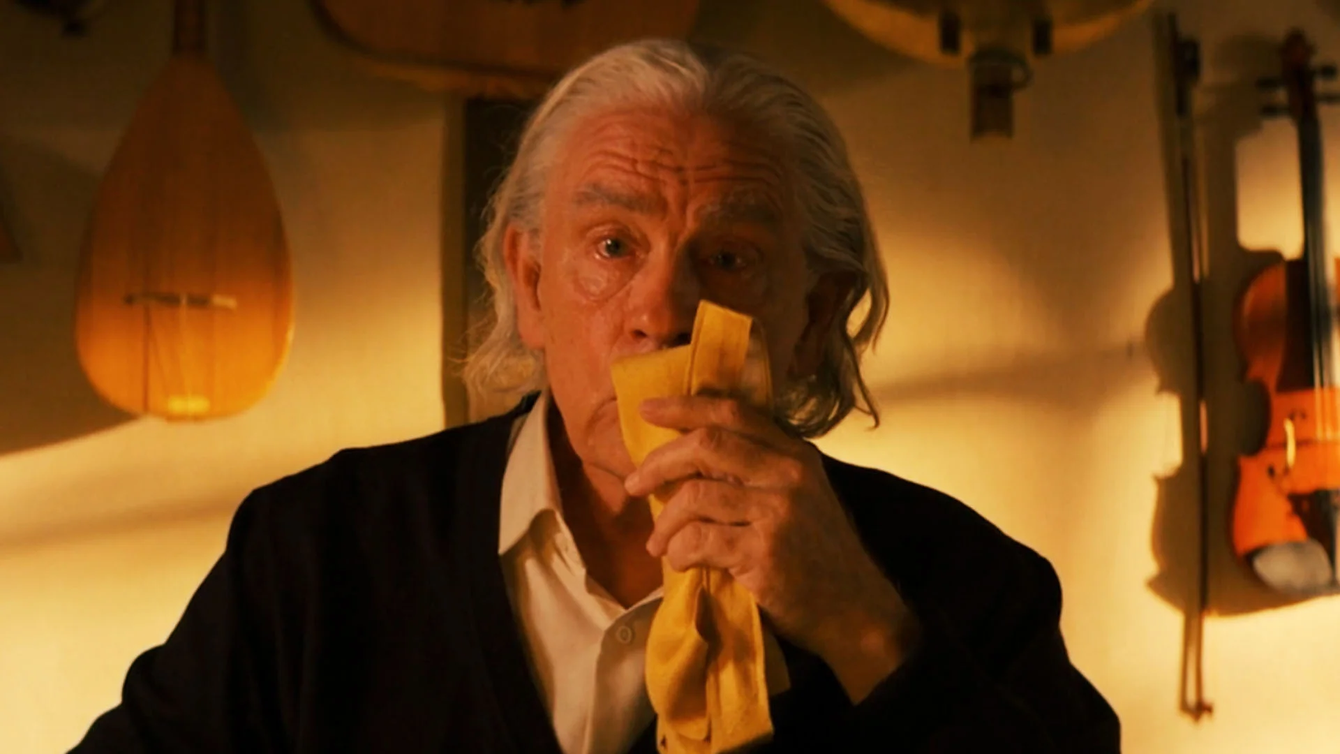 Older Man with White Hair Wipes His Mouth with a Yellow Cloth in a Warm Guitar filled Room