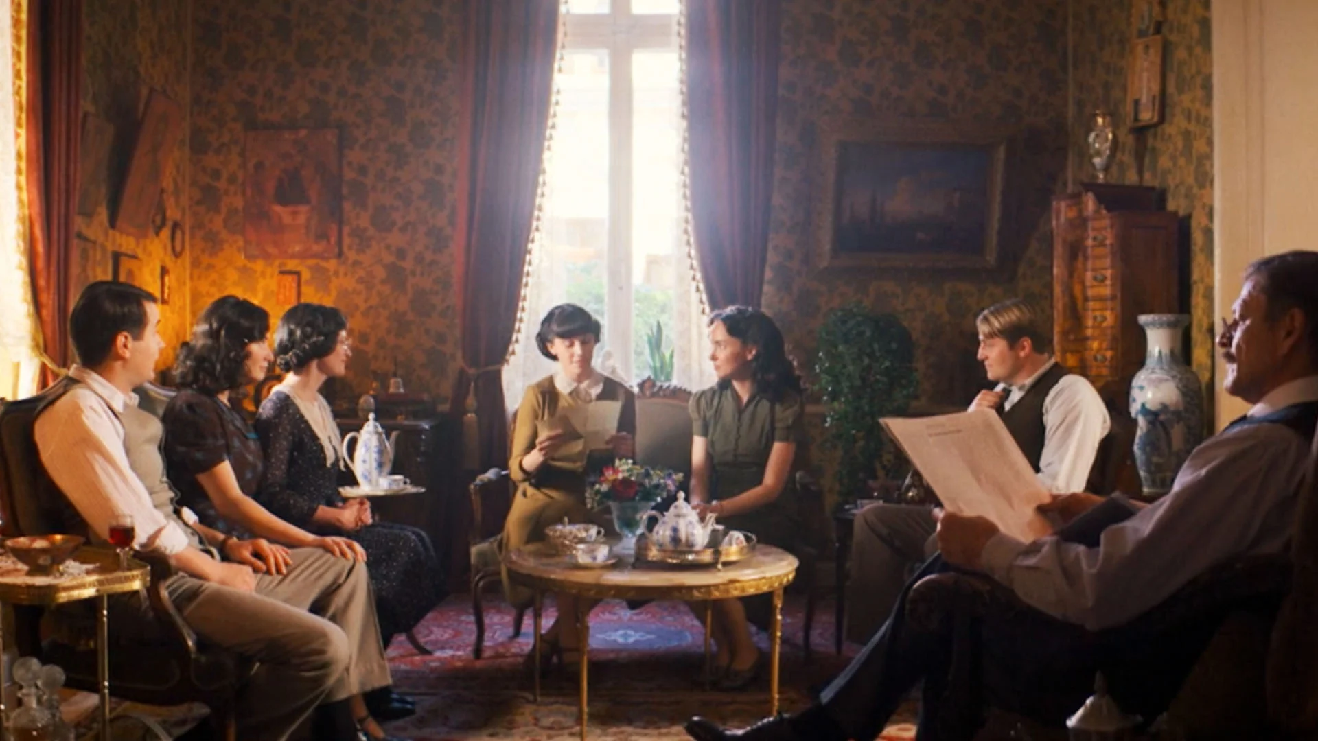 a Group of Men and Women Sit in a Vintage Parlor Around a Coffee Table Reading and Chatting in a Warmly Lit Room