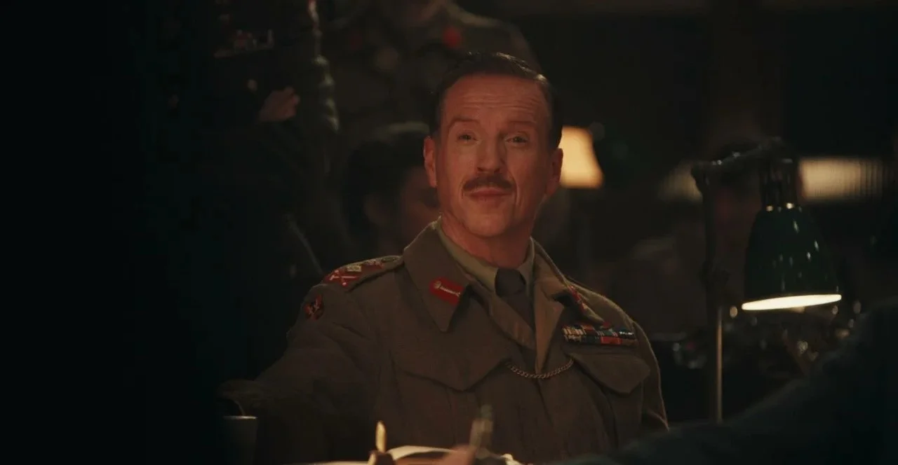 a Mustached Man in an Olive green Military Uniform with Ribbons Sits at a Dimly Lit Table Smiling in a Vintage Dining Room Setting