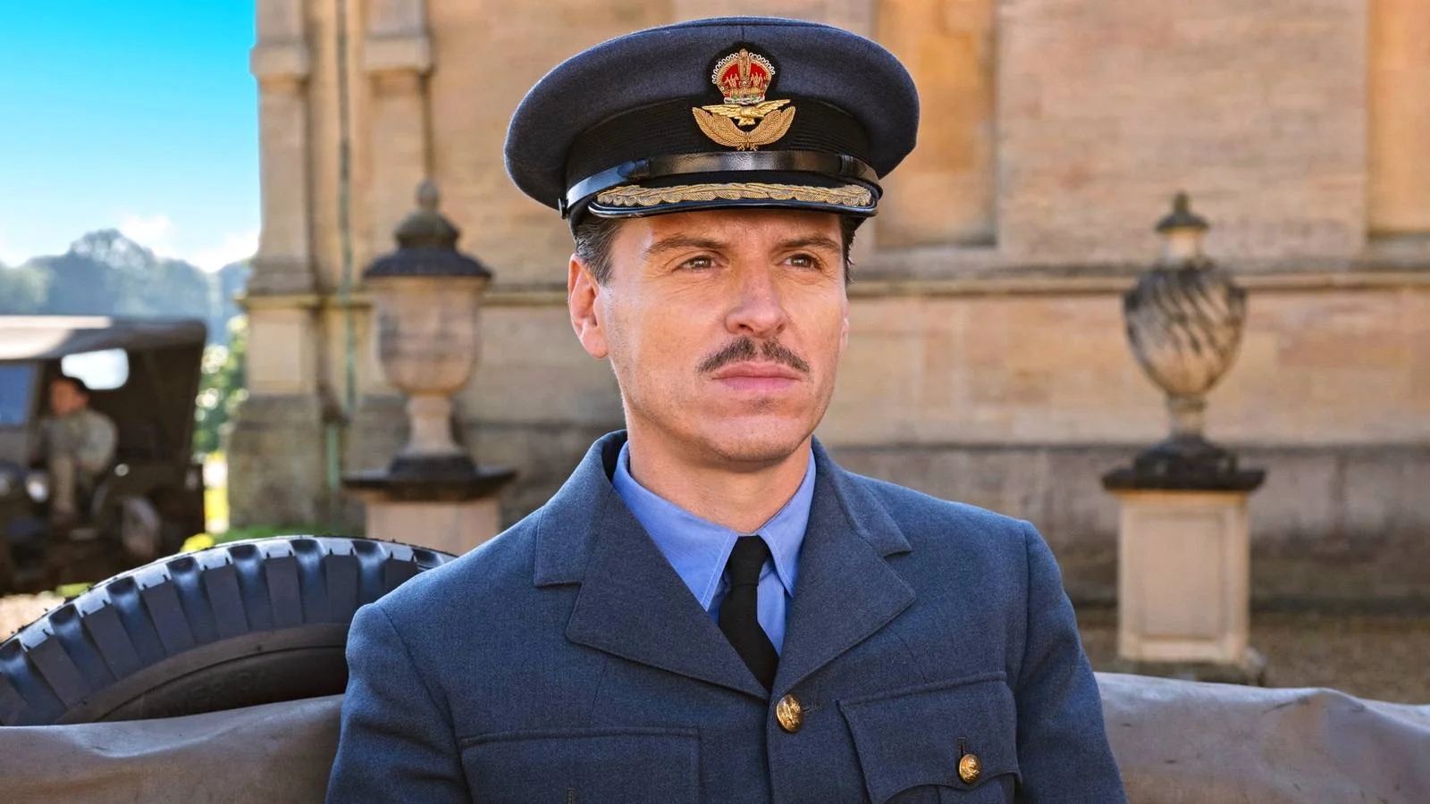 Man in a Vintage Military Uniform blue Jacket and Peaked Cap Seated Outdoors with a Historic Building in the Background
