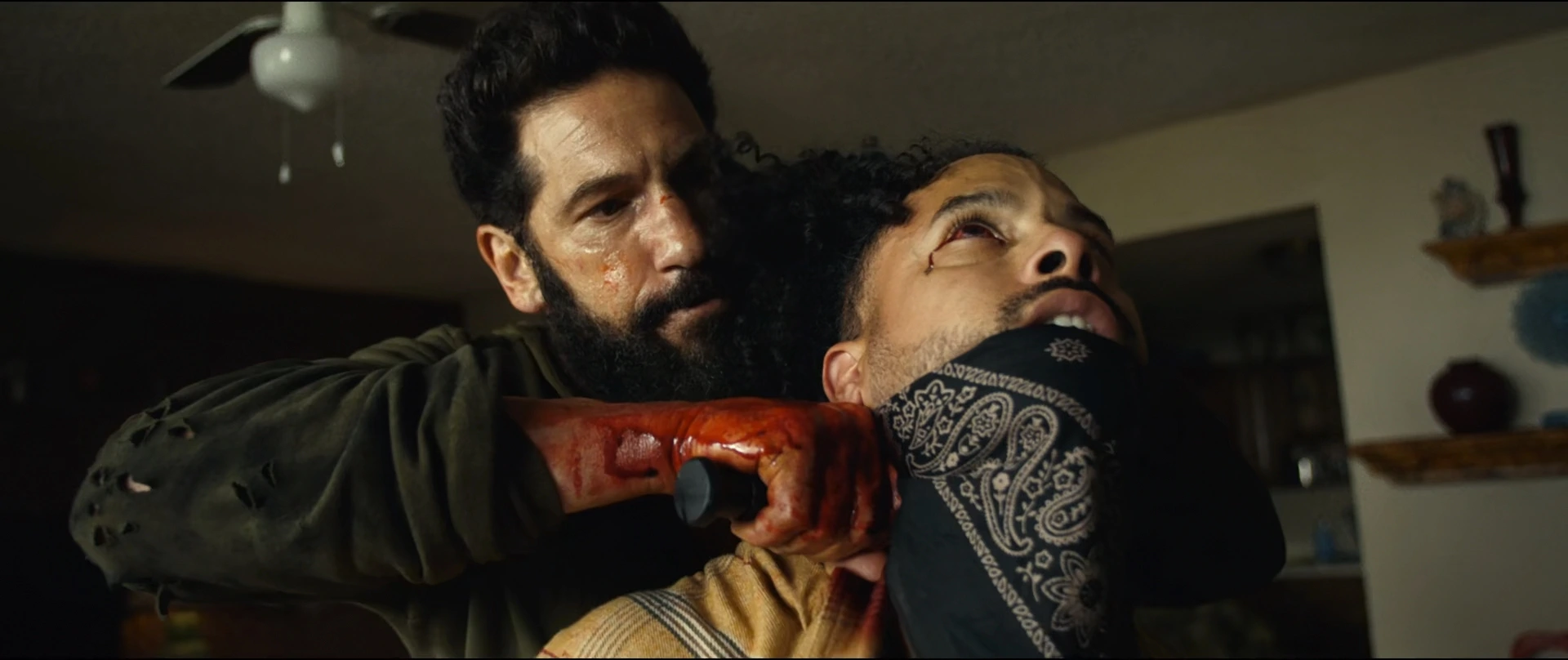 Man with a Beard Grips a Bloodied Knife Against Another Mans Neck in a Dim Living Room
