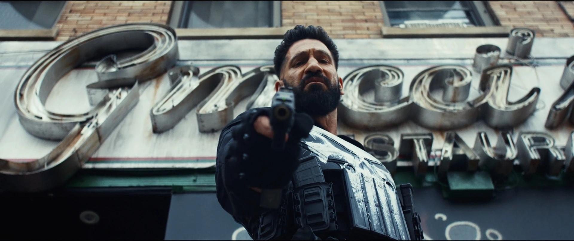Bearded Man in Tactical Gear Points a Handgun at the Viewer in Front of a Neon Storefront Sign