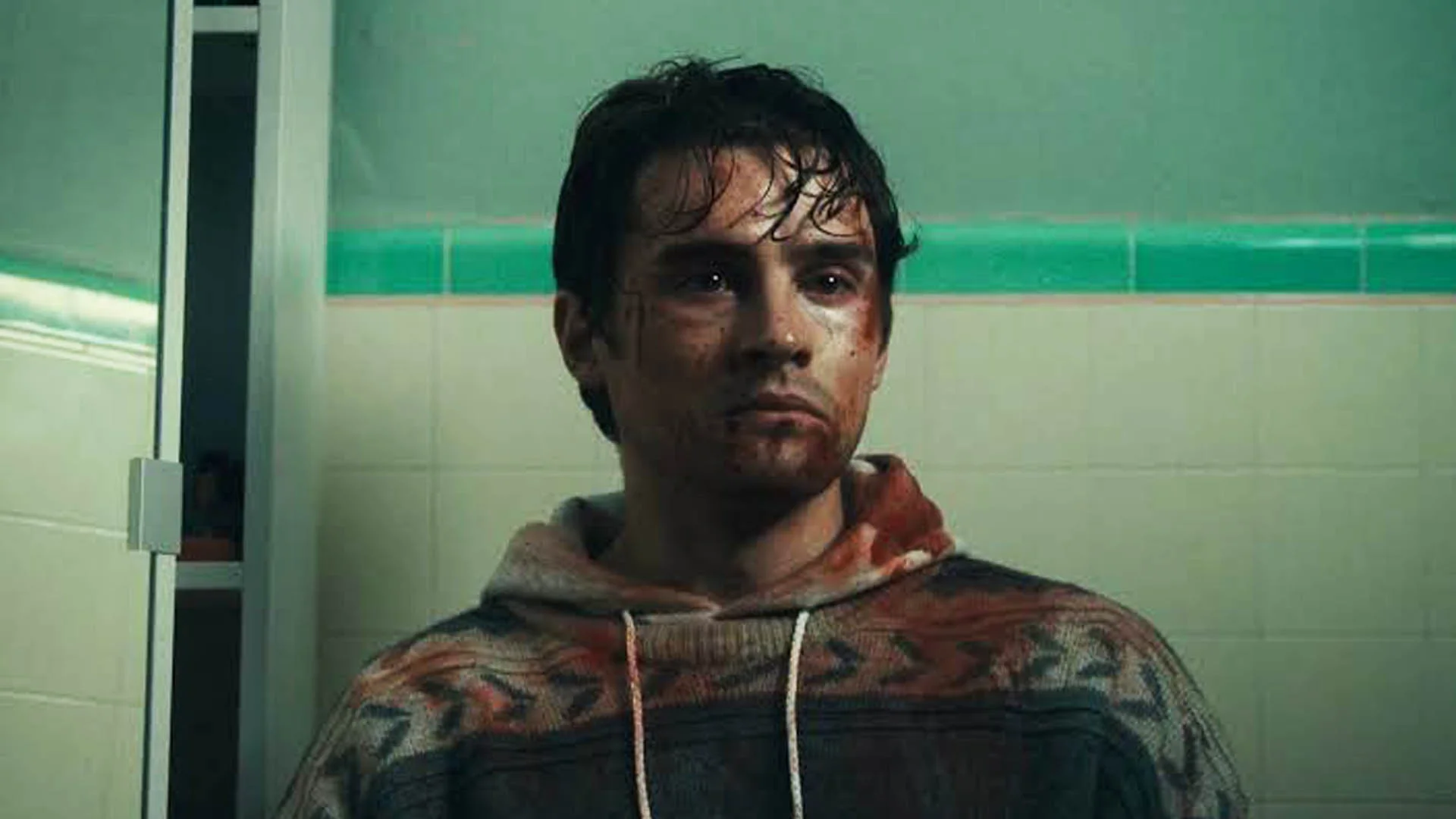 Injured Man with a Bloodied Face Wearing a Hoodie Stands in a Tiled Bathroom with Green Accents Looking off to the Side