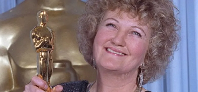 1000051384 Smiling Woman with Curly Hair Holding an Oscar Statuette at an Awards Event in Front of a Blue Backdrop