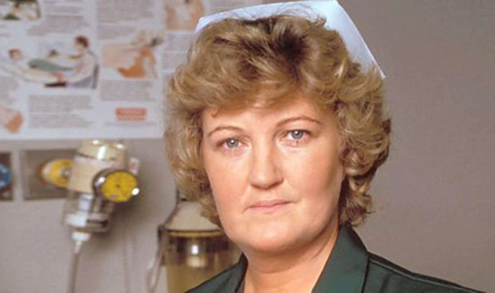 1000051387 Medical Professional with Blond Hair in a Dark Green Uniform Standing in a Clinical Room with Charts and Equipment in the Background