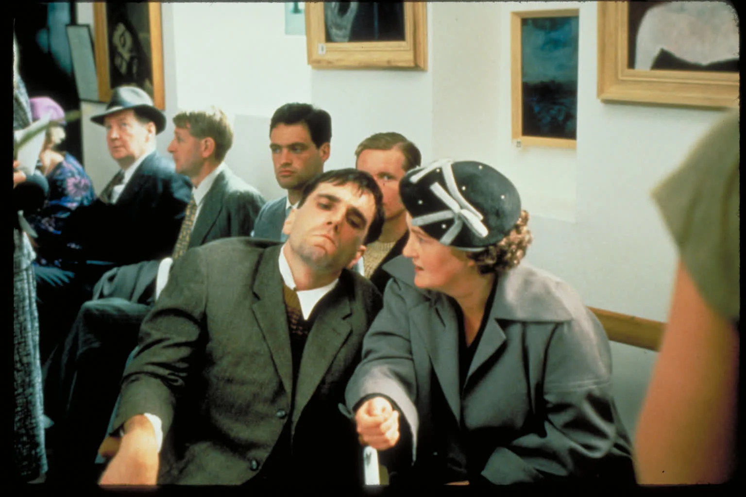 1000051389 a Man in a Suit Leans Forward Beside a Woman in a Gray Coat and Decorative Hat Seated in a Room with Framed Artwork on the Wall and Several Other People in the Background