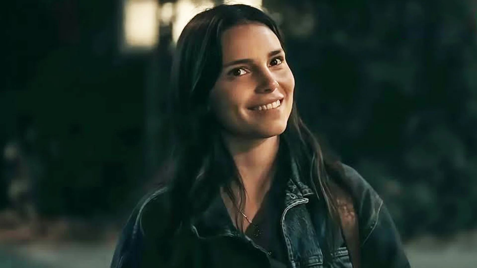 Smiling Woman with Long Dark Hair Wearing a Denim Jacket Outdoors in a Dimly Lit Setting