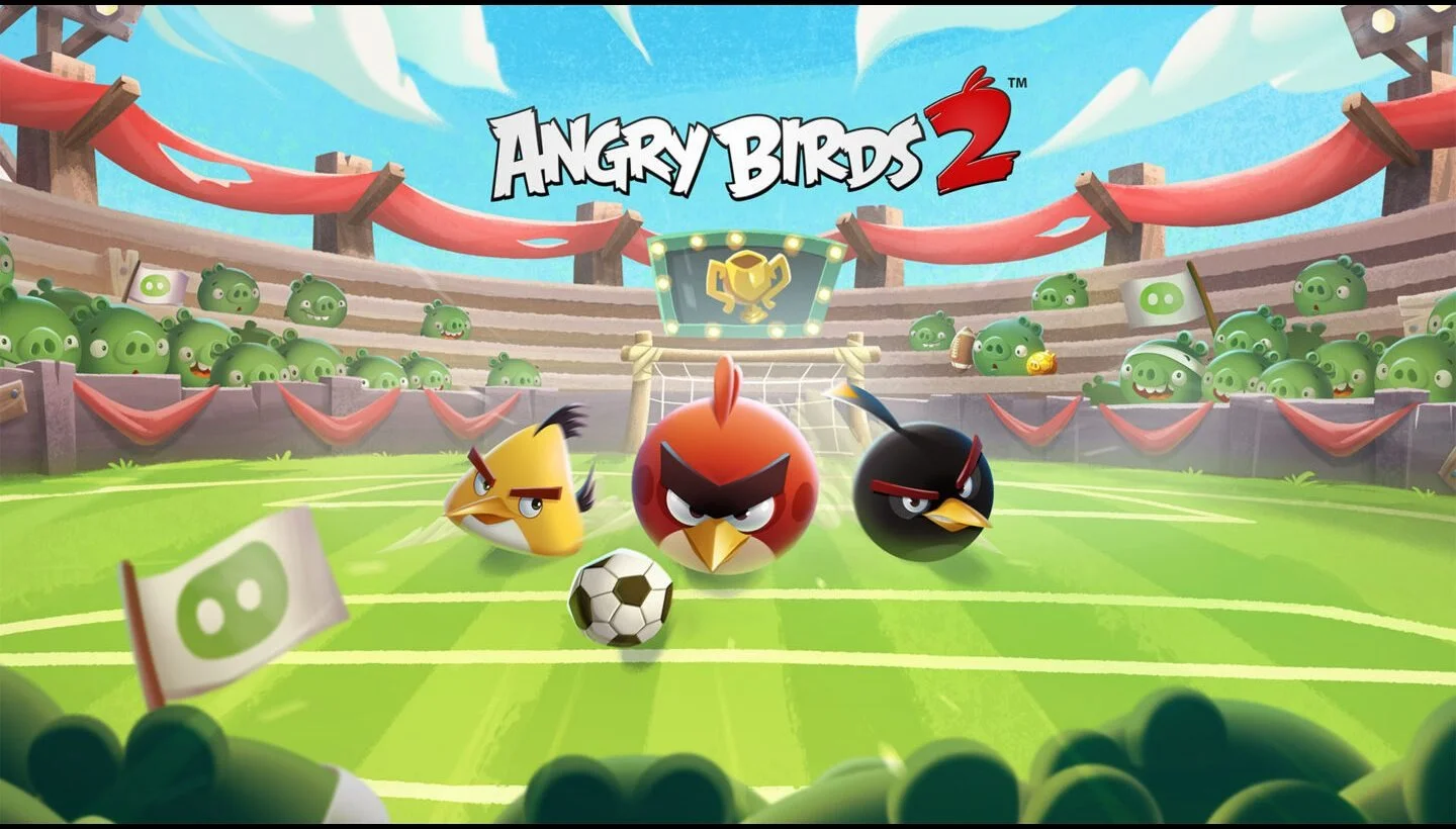 Three Angry Birds red Yellow Black Chase a Soccer Ball on a Bright Stadium Field with Cheering Green Pigs in the Audience Game Logo Above
